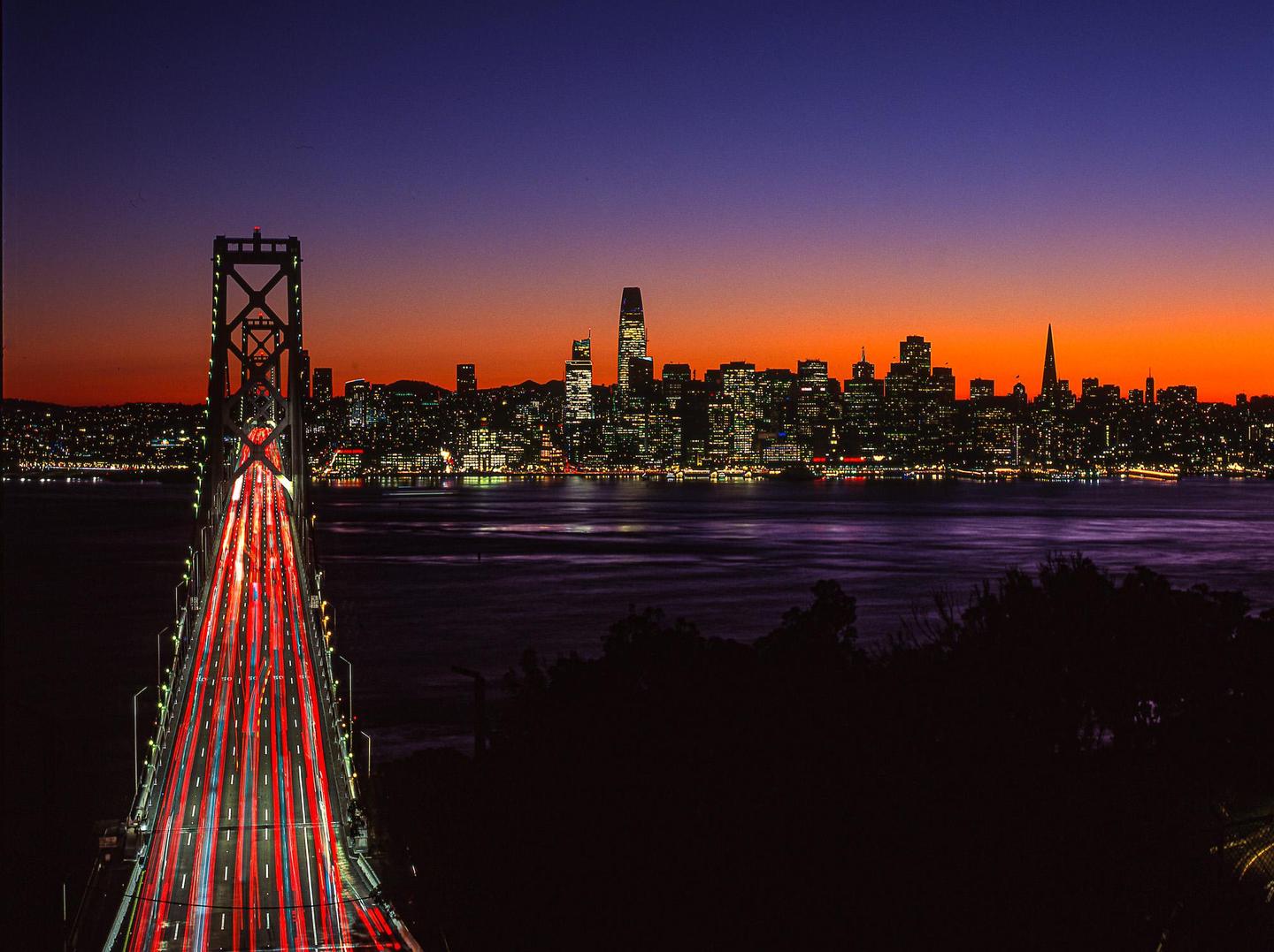 A bridge with light trails from cars leads to a city skyline at dusk.
