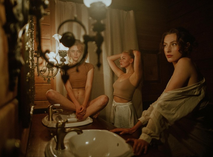 Three women are in a dimly lit bathroom, one sitting on the counter, one fixing her hair, and another leaning on the counter.