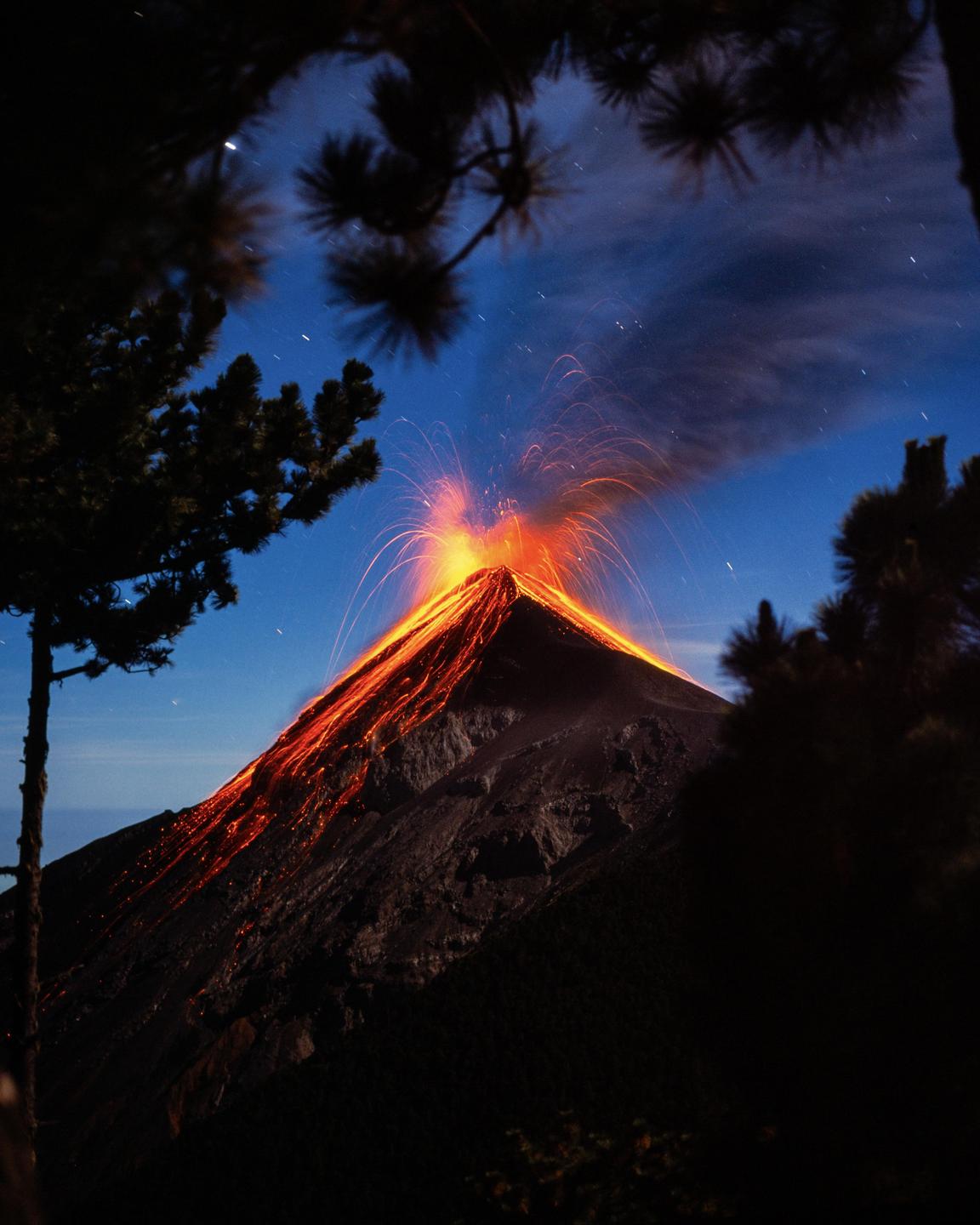 A volcano erupts at night, spewing lava and smoke into the starry sky, framed by silhouetted trees.