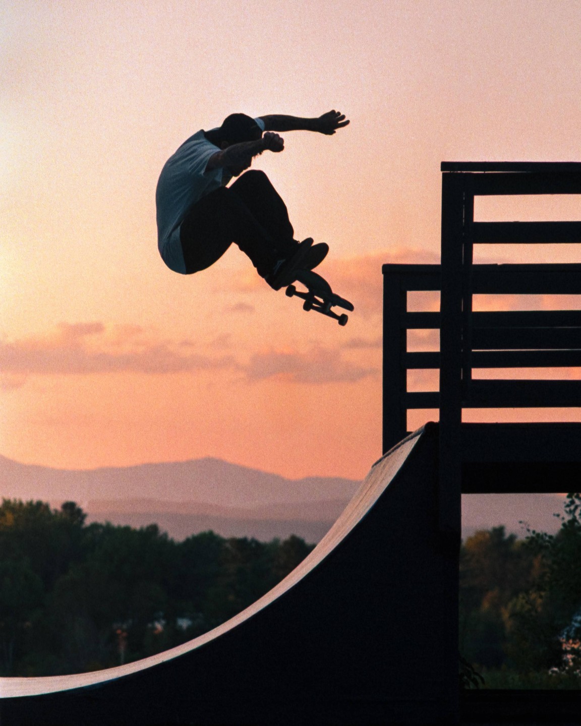 A skateboarder is silhouetted against a colorful sky as he performs a trick over a ramp.