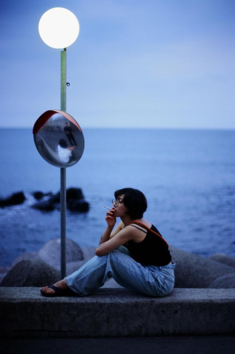 A woman sits on a concrete barrier by the sea, smoking a cigarette, with a convex mirror and a street light behind her.