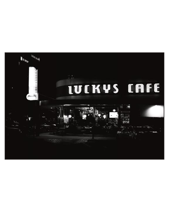 A black and white night street view shows the exterior of Lucky's Cafe with its neon sign illuminated.