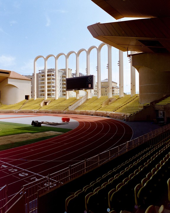A wide view of the Louis II stadium in Monaco shows empty yellow seats, a red running track, and a large scoreboard under a clear sky.