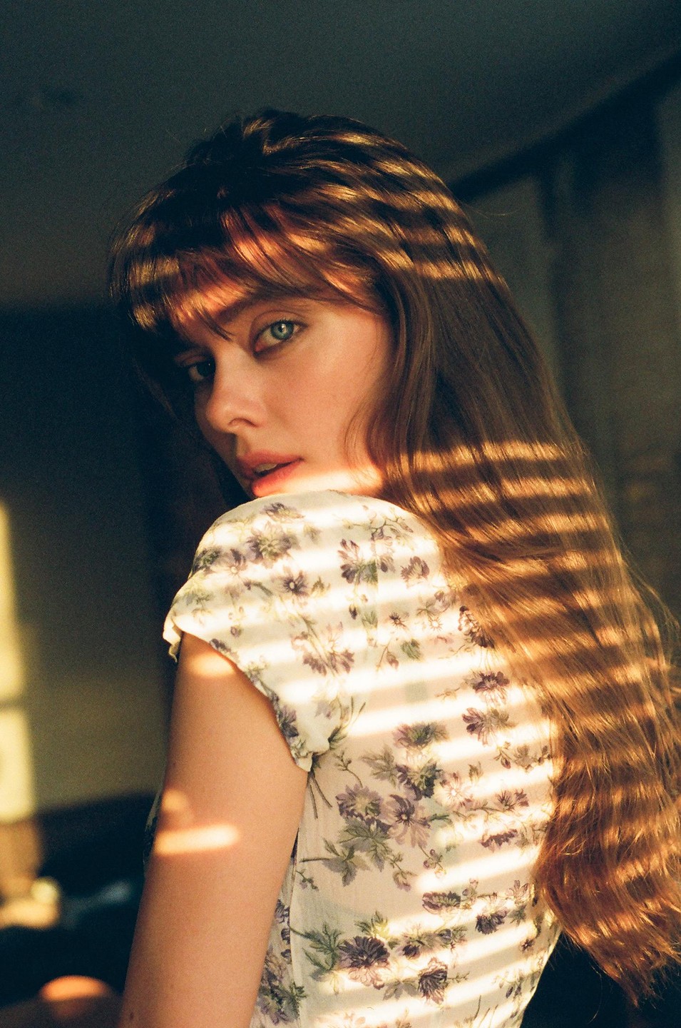 A woman with long hair and bangs looks over her shoulder, illuminated by striped shadows from a window blind.