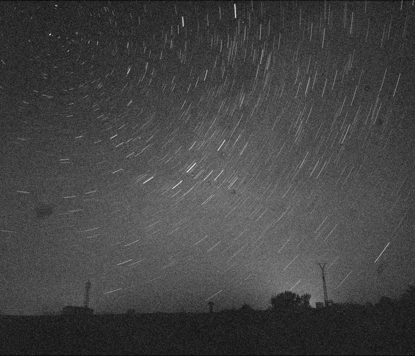 Star trails arc across a dark night sky above a silhouetted landscape with trees and structures.