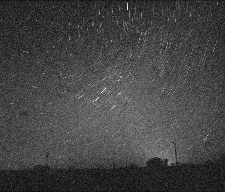 Star trails arc across a dark night sky above a silhouetted landscape with trees and structures.