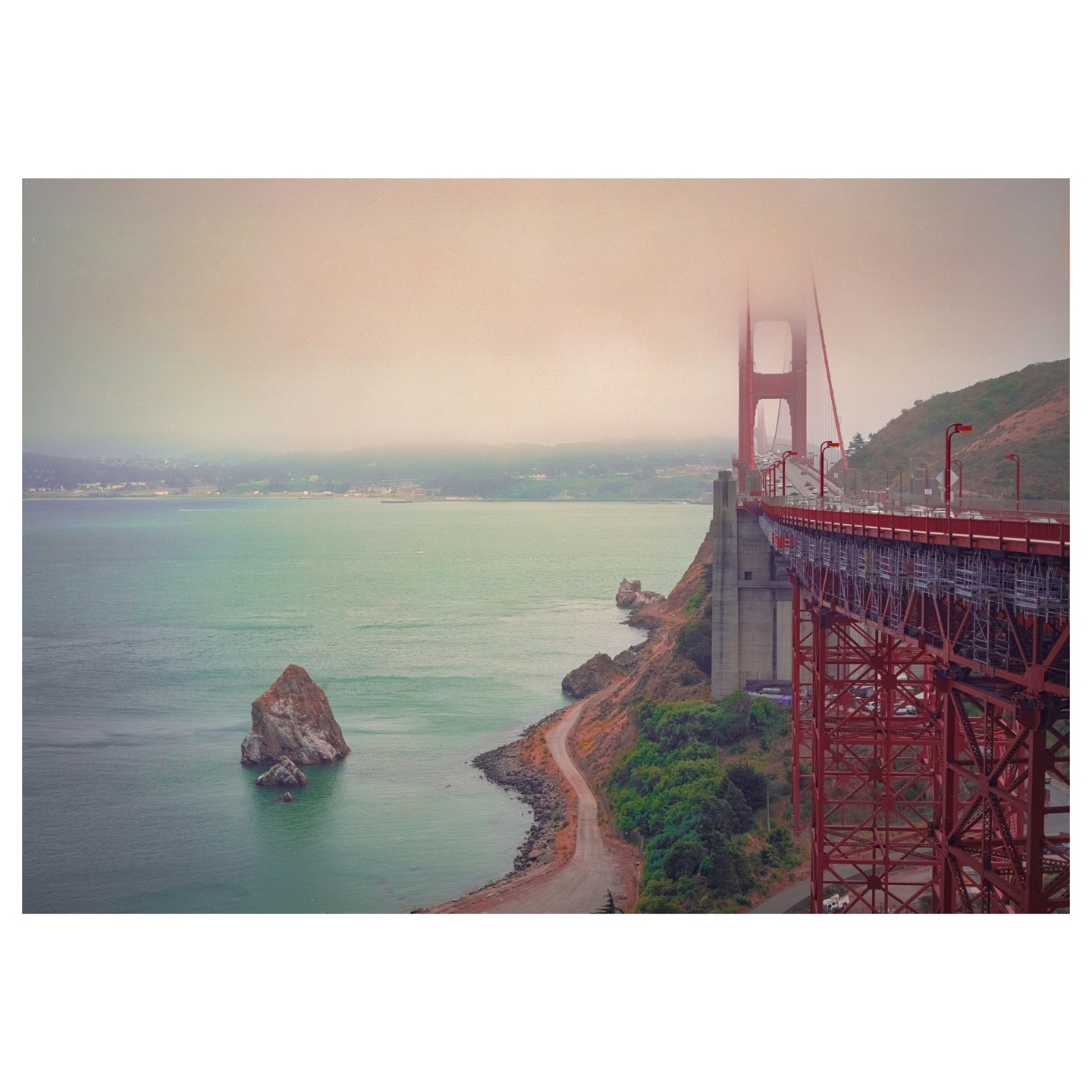 The Golden Gate Bridge extends over a body of water with a rocky shoreline and a single large rock in the water, all under a hazy sky.