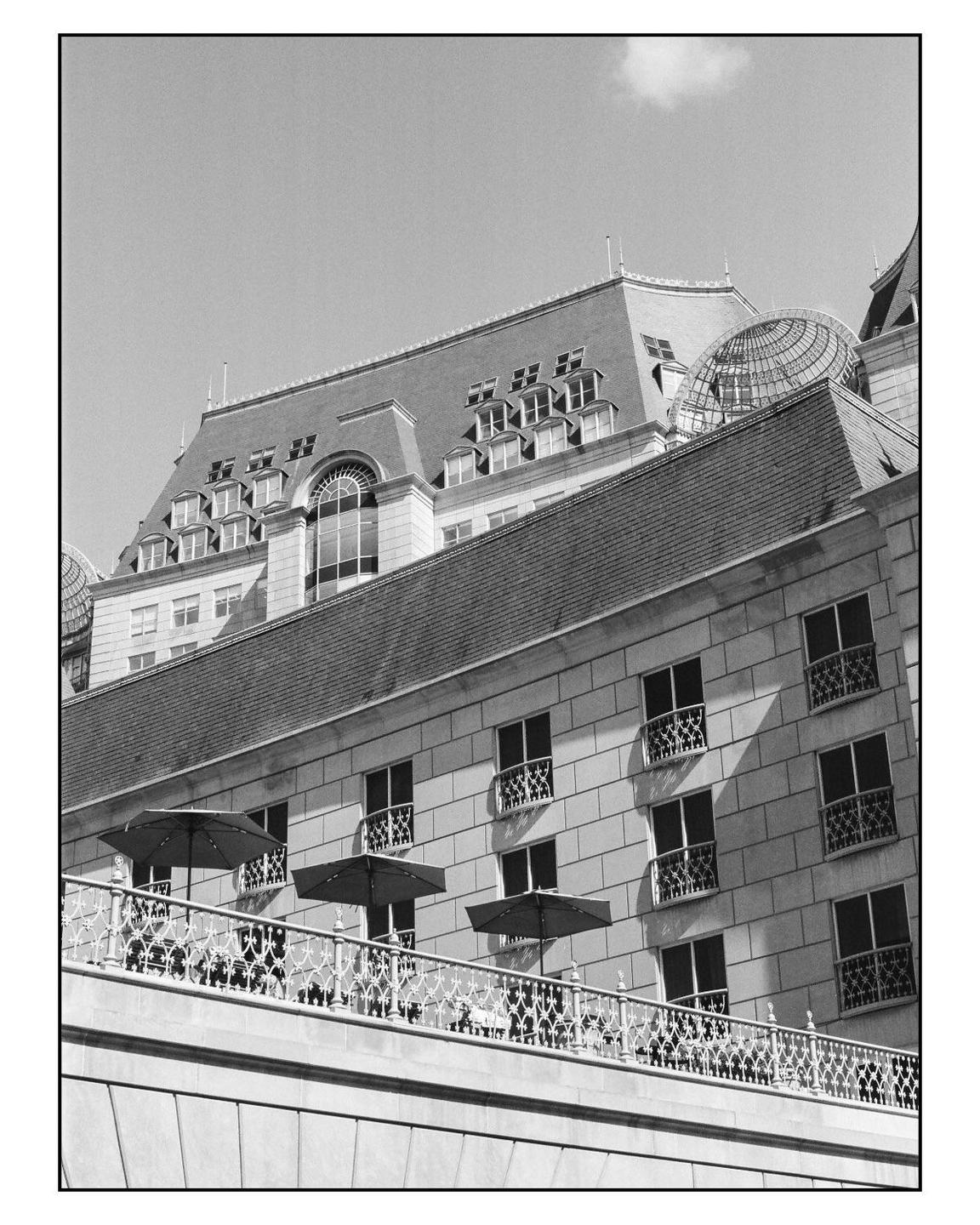A multi-story building with a mansard roof and a balcony with umbrellas is shown from a low angle.