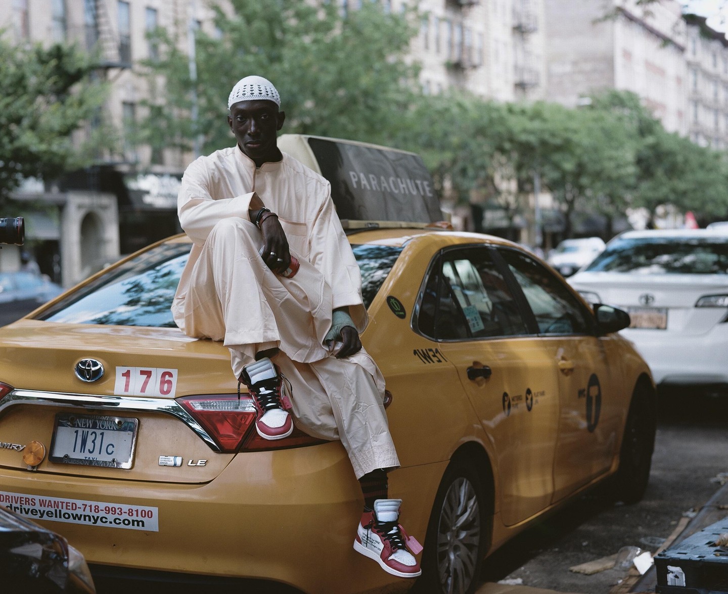 A man wearing a white kufi and light-colored clothing sits on the trunk of a yellow taxi cab in a city.