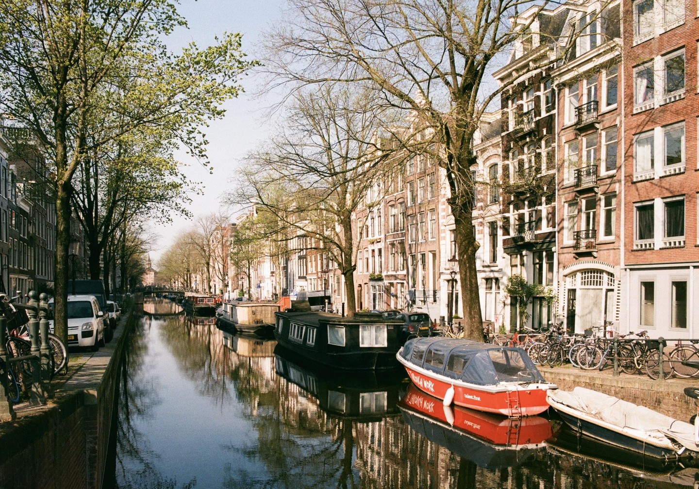 A canal with boats and houseboats is lined with trees and historic buildings in Amsterdam.