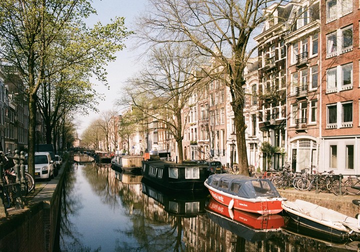 A canal with boats and houseboats is lined with trees and historic buildings in Amsterdam.