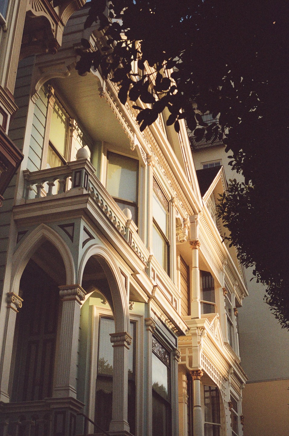 Sunlight illuminates the ornate architectural details of a Victorian house, partially obscured by dark foliage.