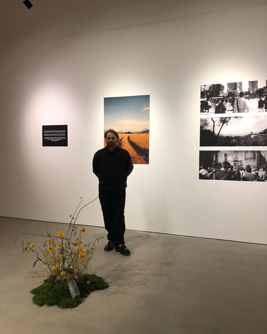 A man stands in an exhibition space next to a display of photographs and a floral arrangement.