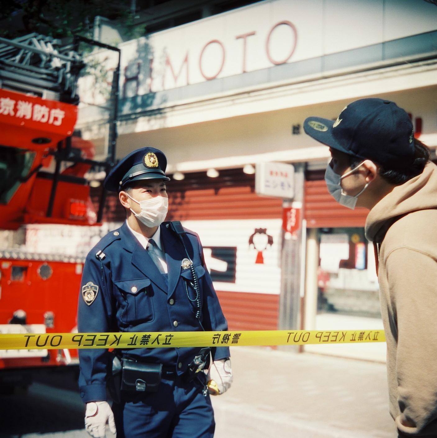 A police officer in uniform and a man in a baseball cap stand on a street separated by yellow tape.