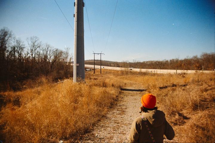 A person in an orange hat walks along a dirt path next to a field of dry grass with a highway and trees in the distance.