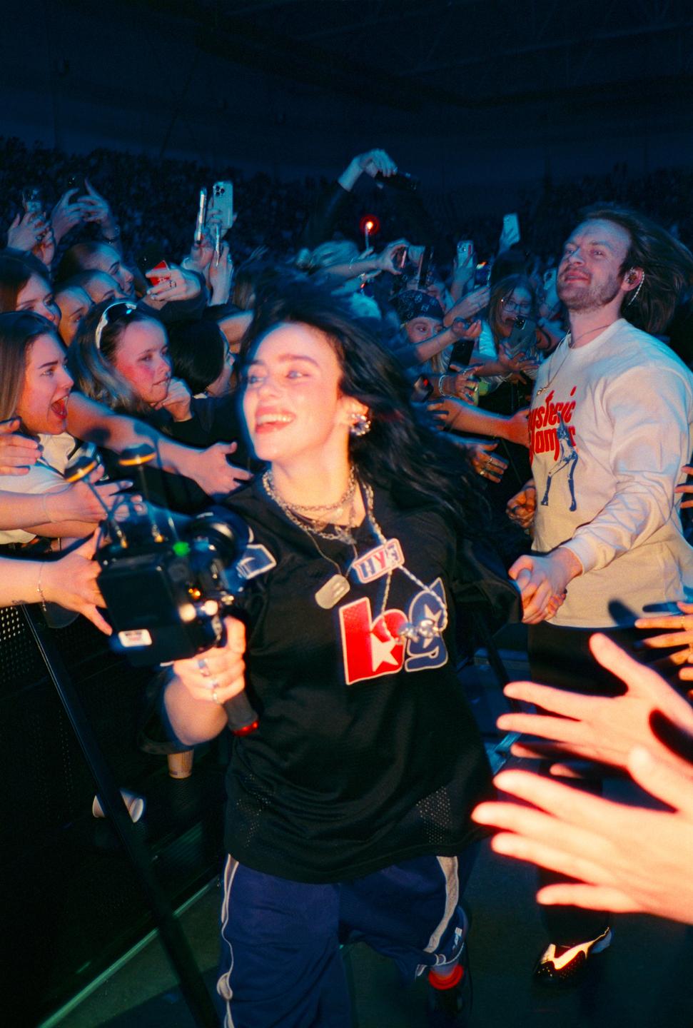 A woman with dark hair smiles while holding a camera and walking through a crowd of people.