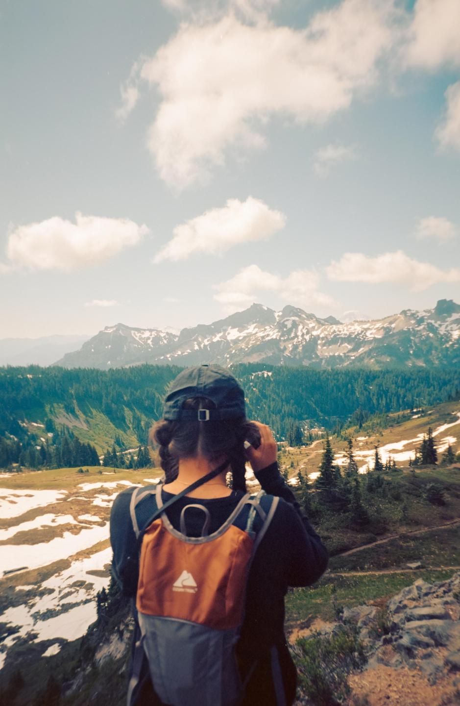 A person with a backpack and a baseball cap looks out at a mountain range with snow and a forest.