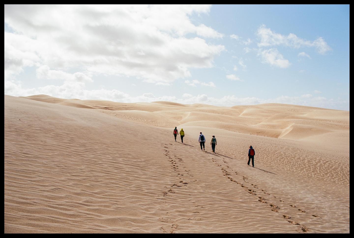 Five people walk in a line across a vast desert landscape under a partly cloudy sky.