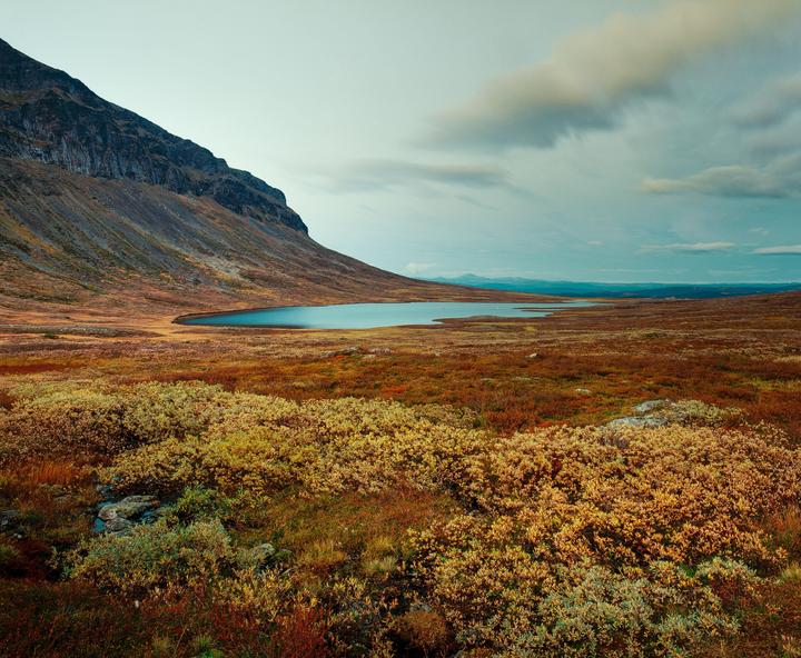A body of water sits at the base of a mountain in a vast, colorful landscape under a cloudy sky.