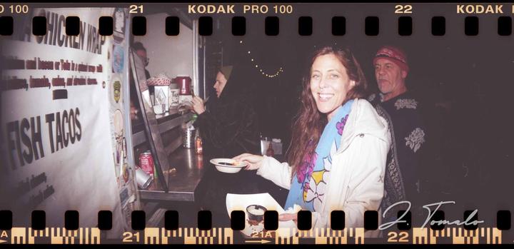 A woman smiles at the camera while holding a small bowl of food at a food truck at night.