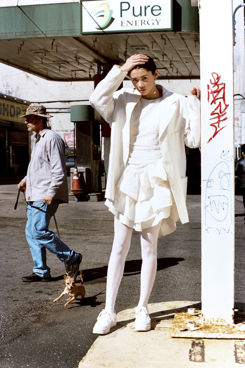 A woman in white clothing stands next to a pillar with graffiti, while a man walks a small dog on a leash in the background.