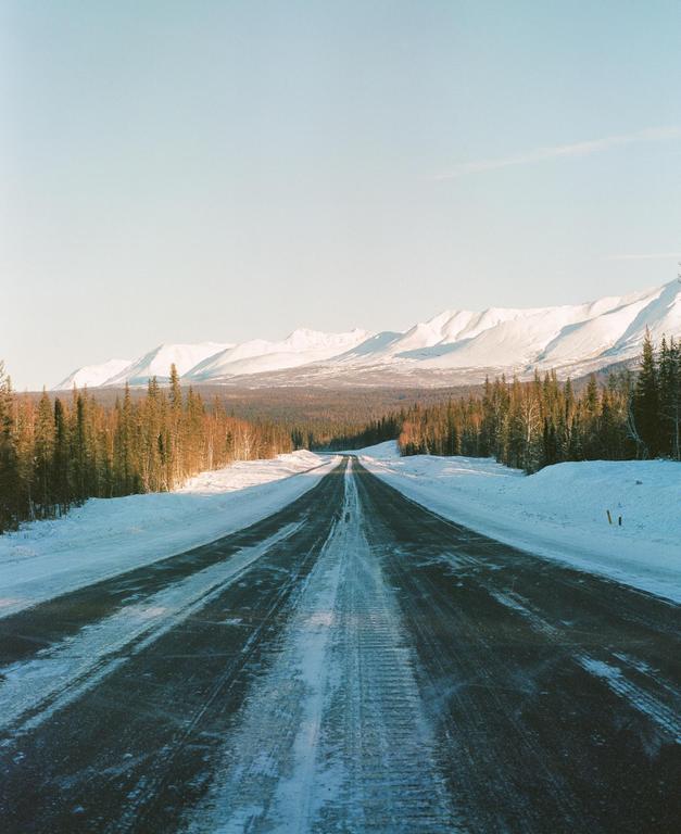 A long, snow-covered road stretches into the distance, flanked by evergreen trees and snow-capped mountains under a clear sky.
