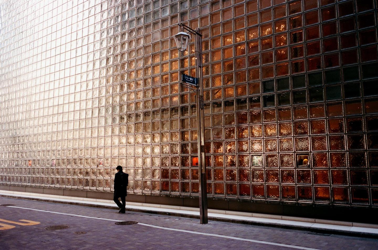A person walks along a street next to a building with a glass block facade.