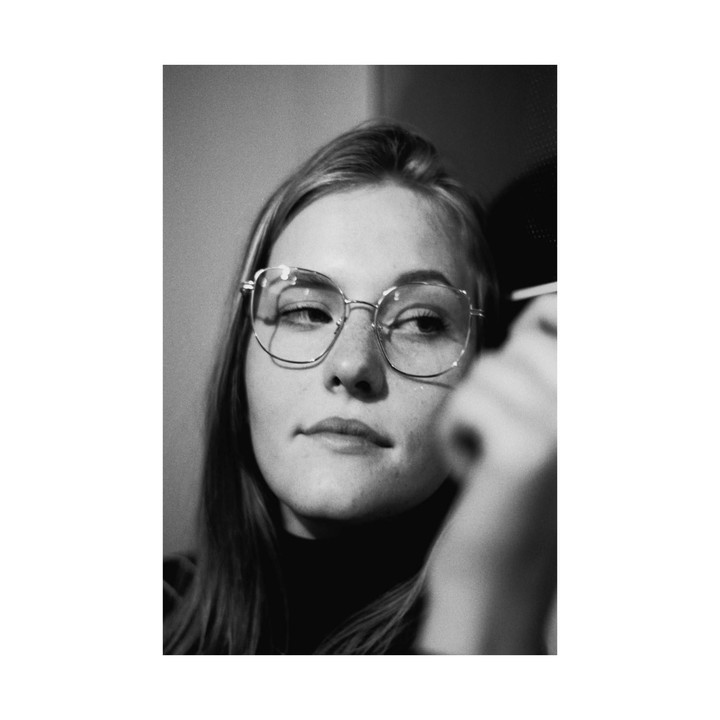 A woman wearing glasses looks to the side while holding a small stick.