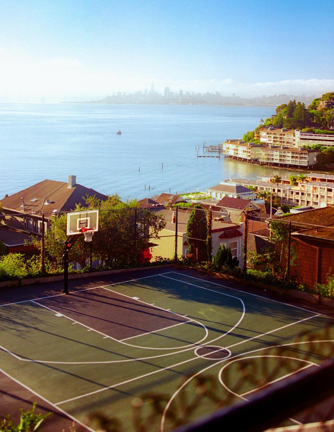 A basketball court overlooks a body of water with a distant city skyline and houses built into a hillside.
