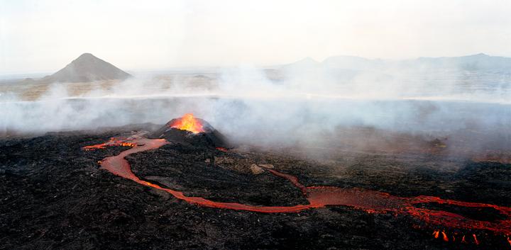 A volcano erupts, with lava flowing across a dark, rocky landscape and smoke rising into a hazy sky.