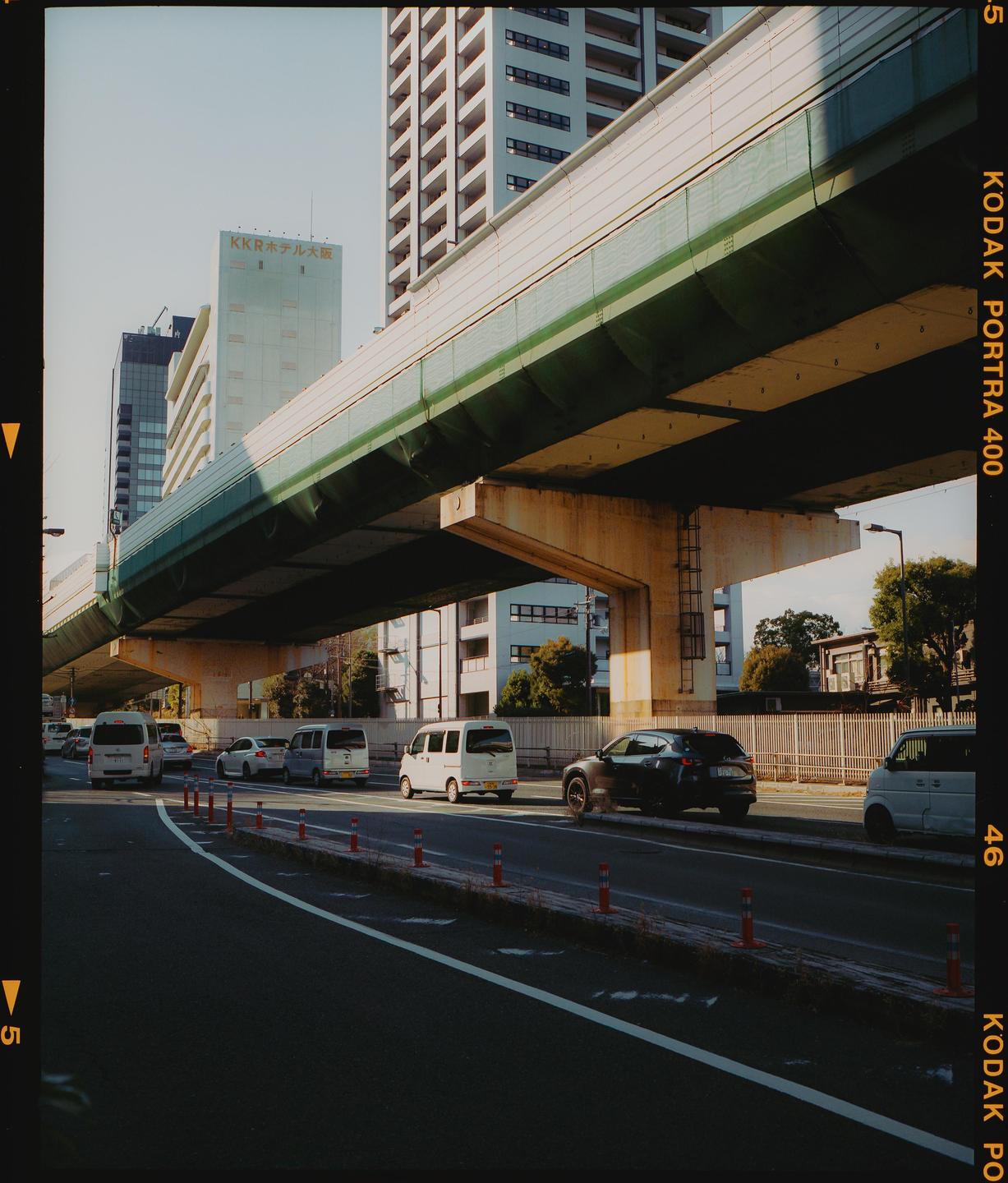 A street scene in Osaka, Japan, shows cars driving under a large concrete overpass with tall buildings in the background.