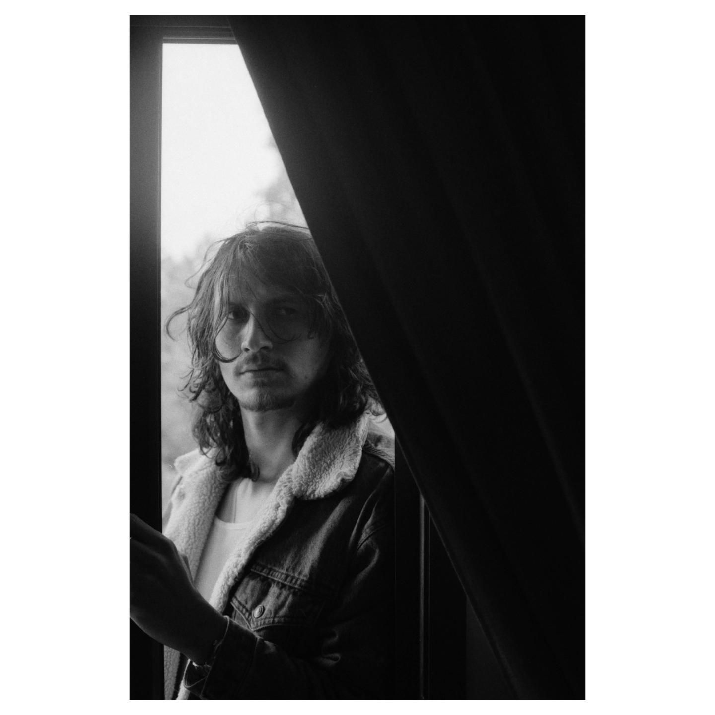 A man with long hair and a denim jacket looks out a window, partially obscured by a dark curtain.