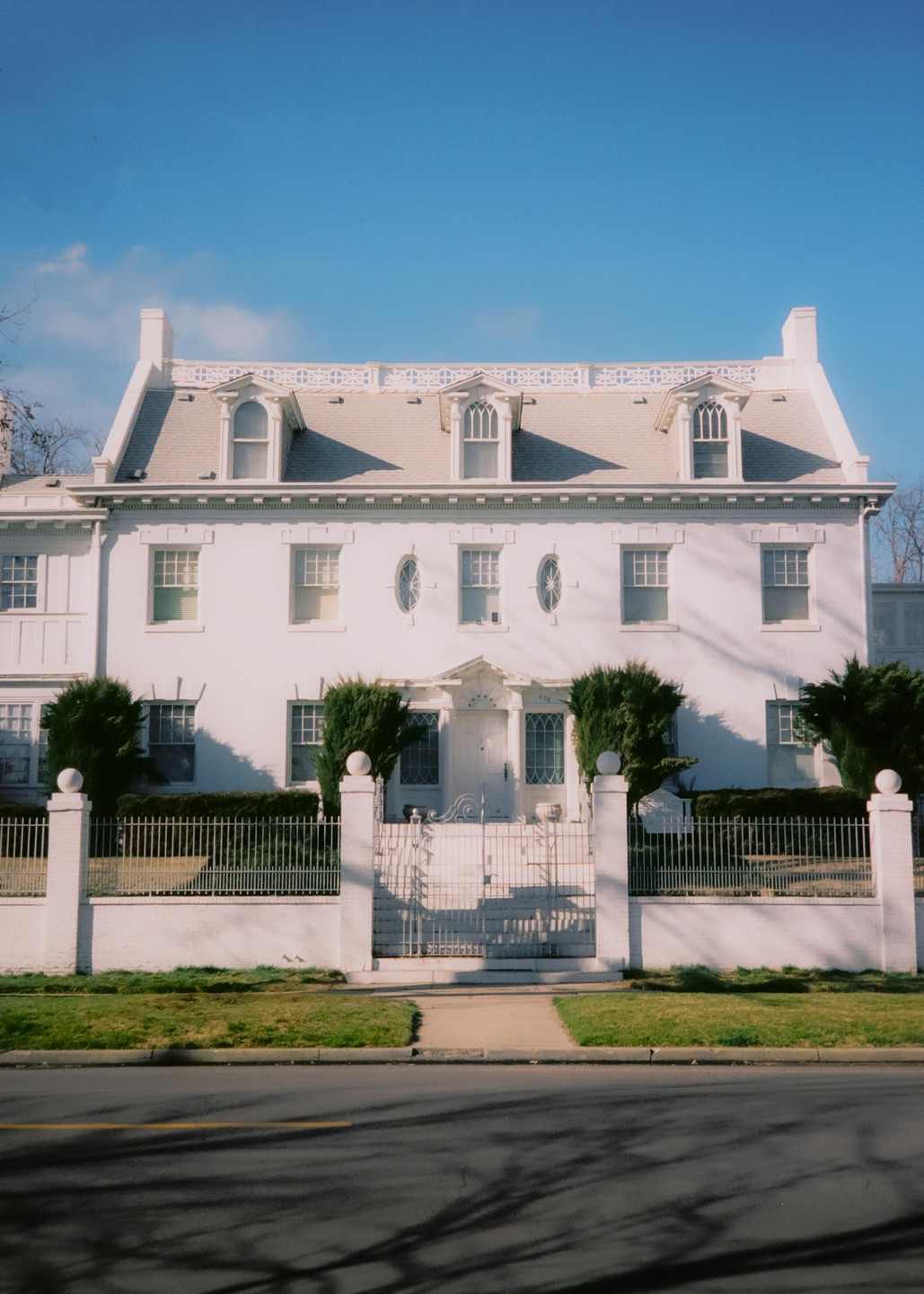 A large white house with a gray roof and dormer windows stands behind a white fence and gate, with a street in the foreground.