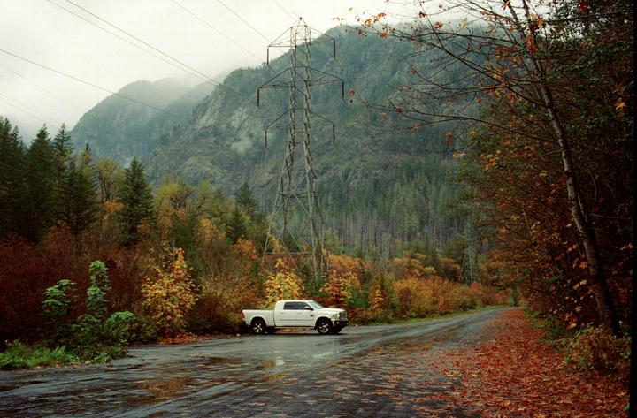 A white pickup truck is parked on a wet road with autumn foliage and a power line tower in the background.