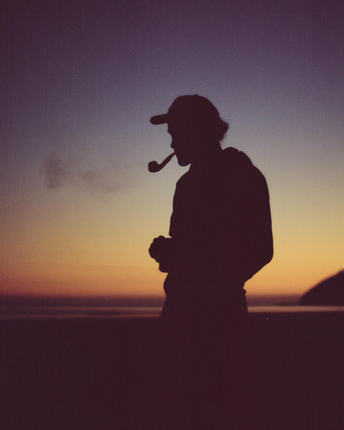 A man in silhouette smokes a pipe on a beach at sunset.