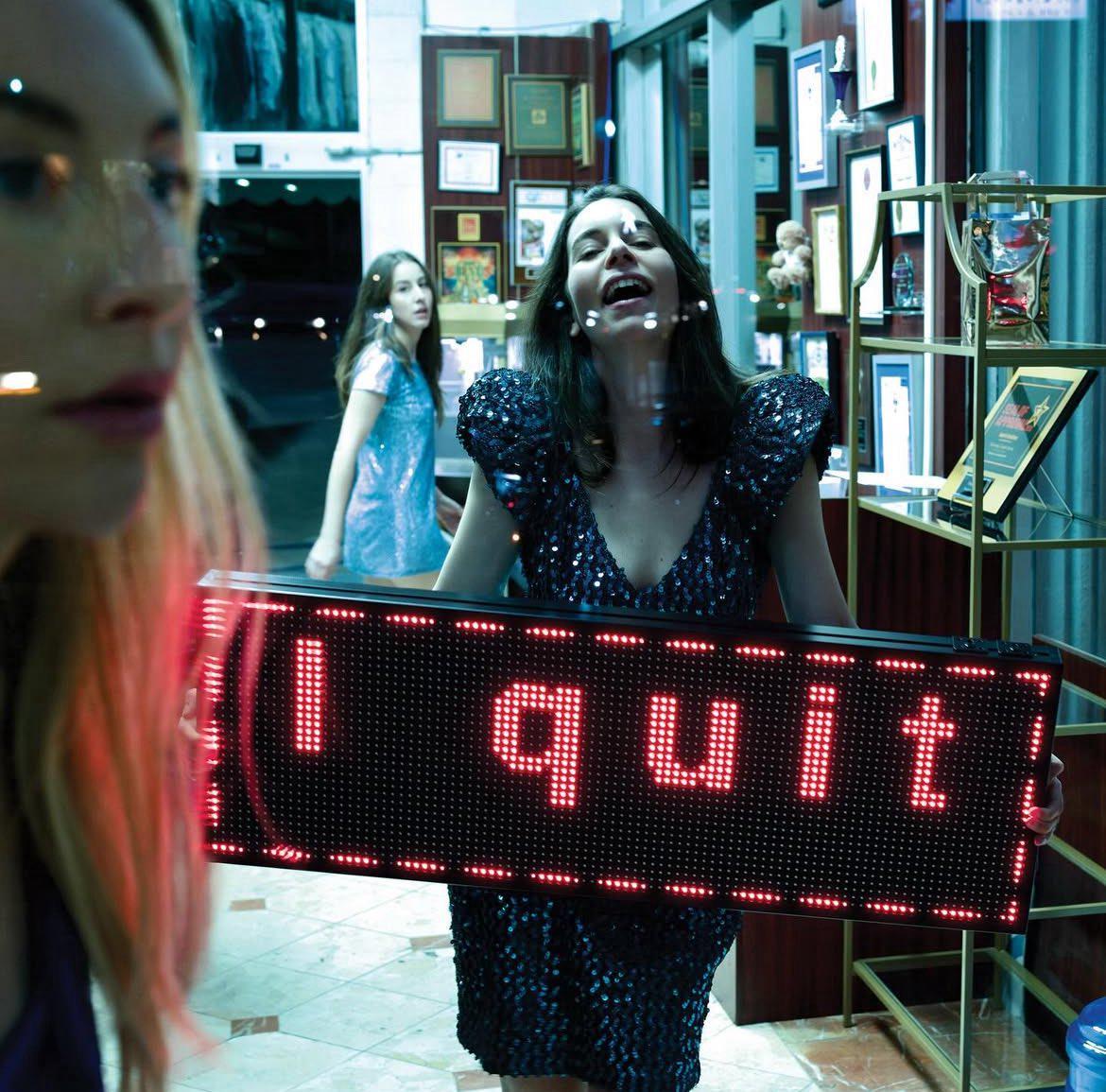 A woman in a sequined dress holds a glowing red sign that says "I quit" while another woman looks on in the background.