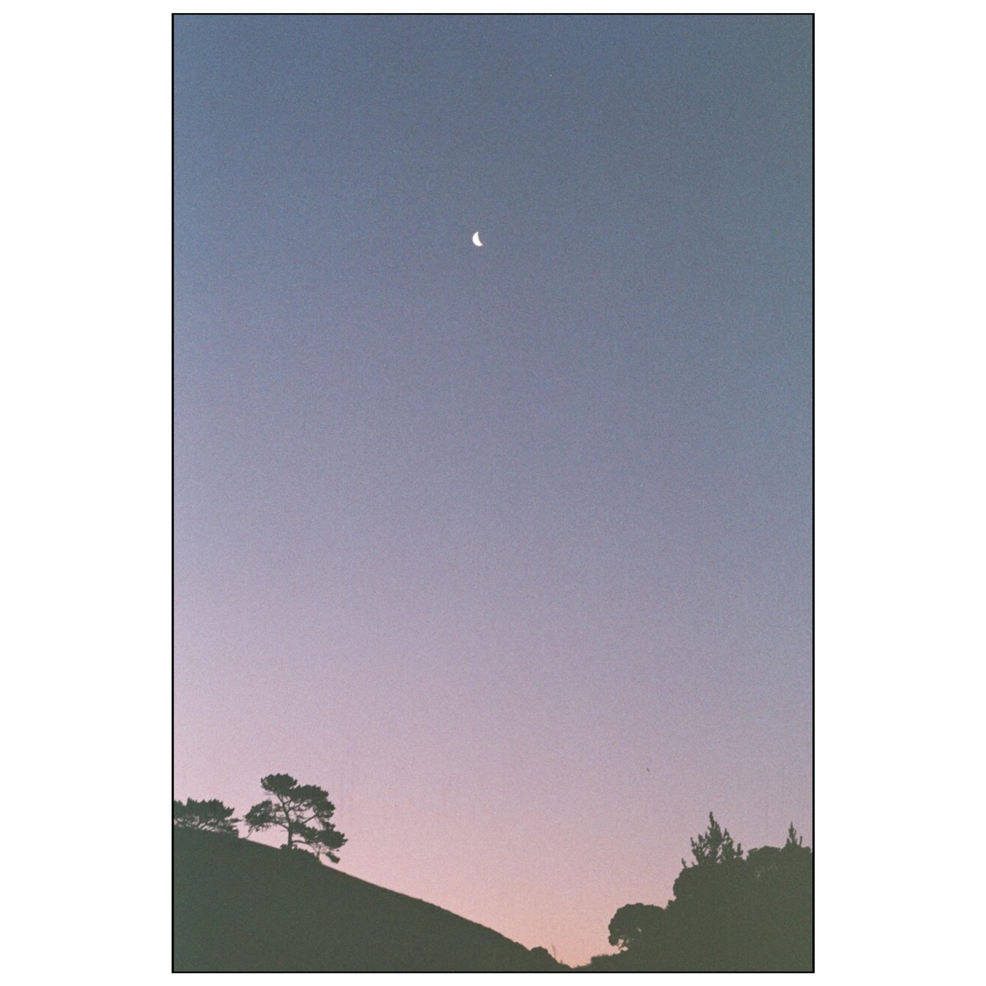 A crescent moon hangs in a gradient sky above a silhouetted hillside with trees.