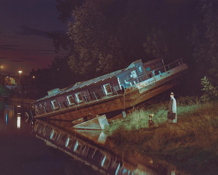 A man in a trench coat and hat stands with a dog on a grassy bank next to a partially submerged, derelict boat at night.