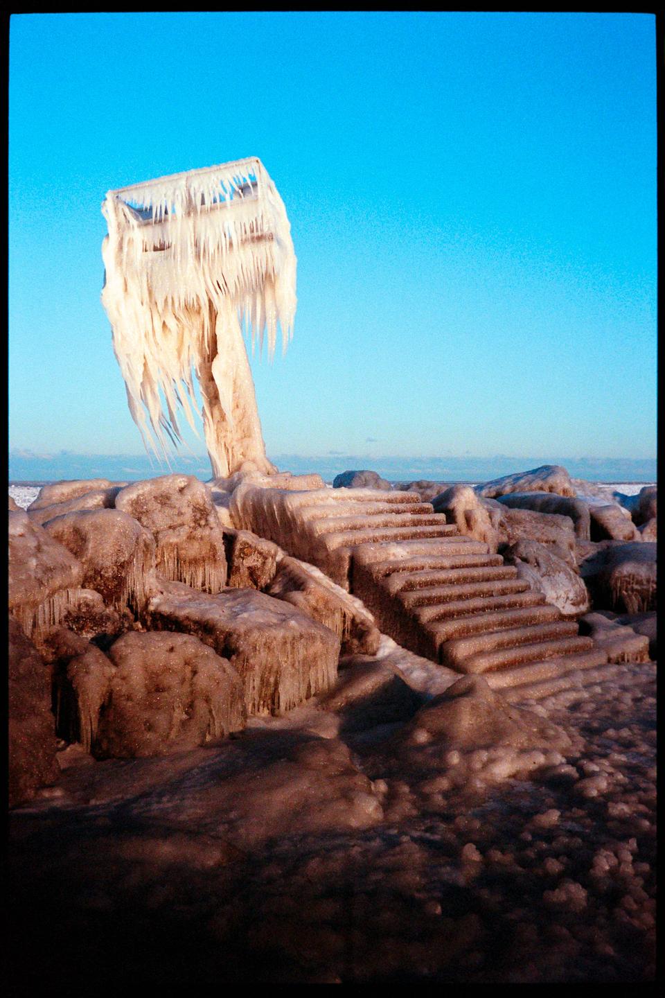 A structure covered in thick ice and icicles stands next to ice-covered steps and rocks under a clear blue sky.