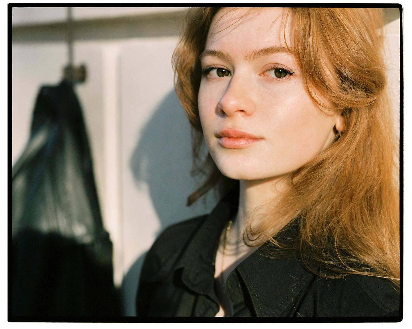 A woman with long red hair and a black shirt looks directly at the viewer, with a shadow on the wall behind her.