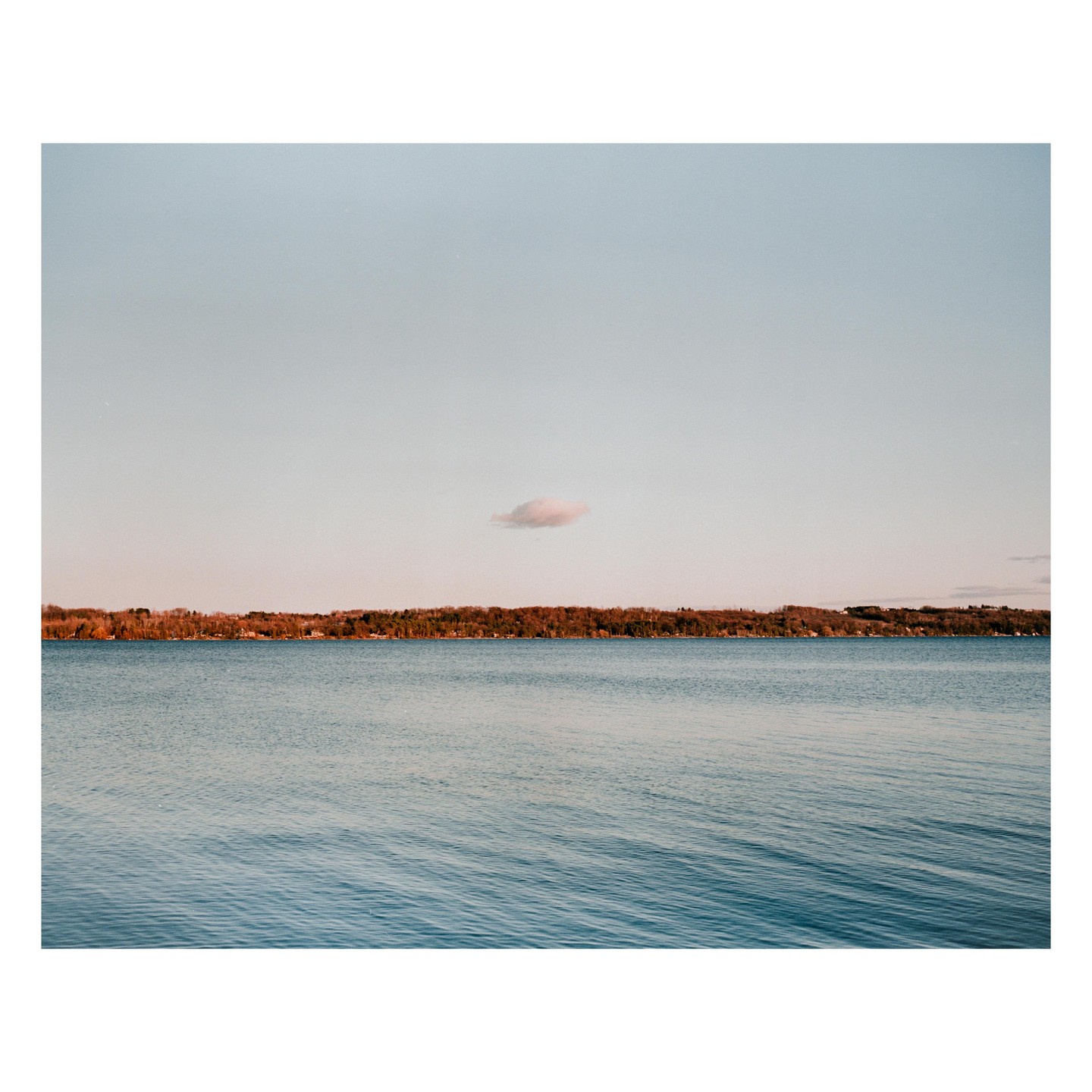 A single cloud floats above a body of water with a tree-lined shore under a pale sky.