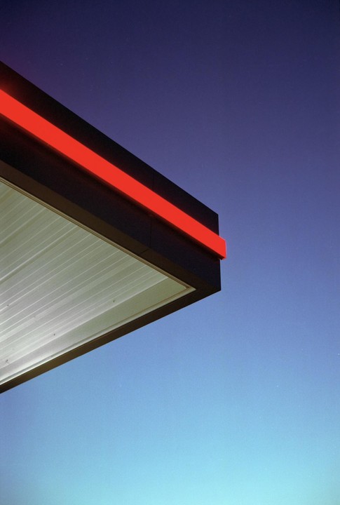 A gas station canopy with a red stripe is visible against a blue sky at night.