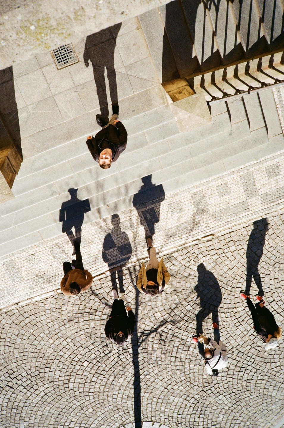 A high-angle shot shows several people and their shadows on a cobblestone street and stone steps.