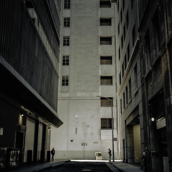Two men stand in a narrow alley between tall buildings, with one man on the left and another on the right.