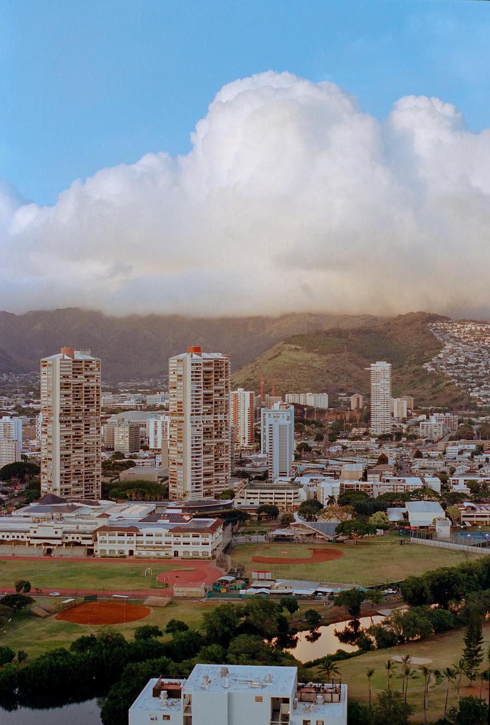 A city with tall buildings and a baseball field is nestled at the base of a mountain range under a cloudy sky.