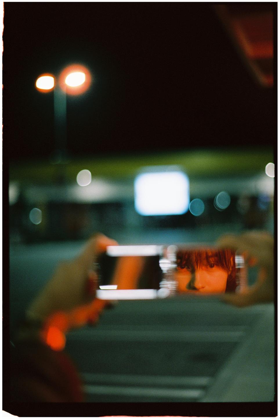 A person holds a phone displaying a reflection of a woman's face with bangs, against a blurred background of streetlights at night.