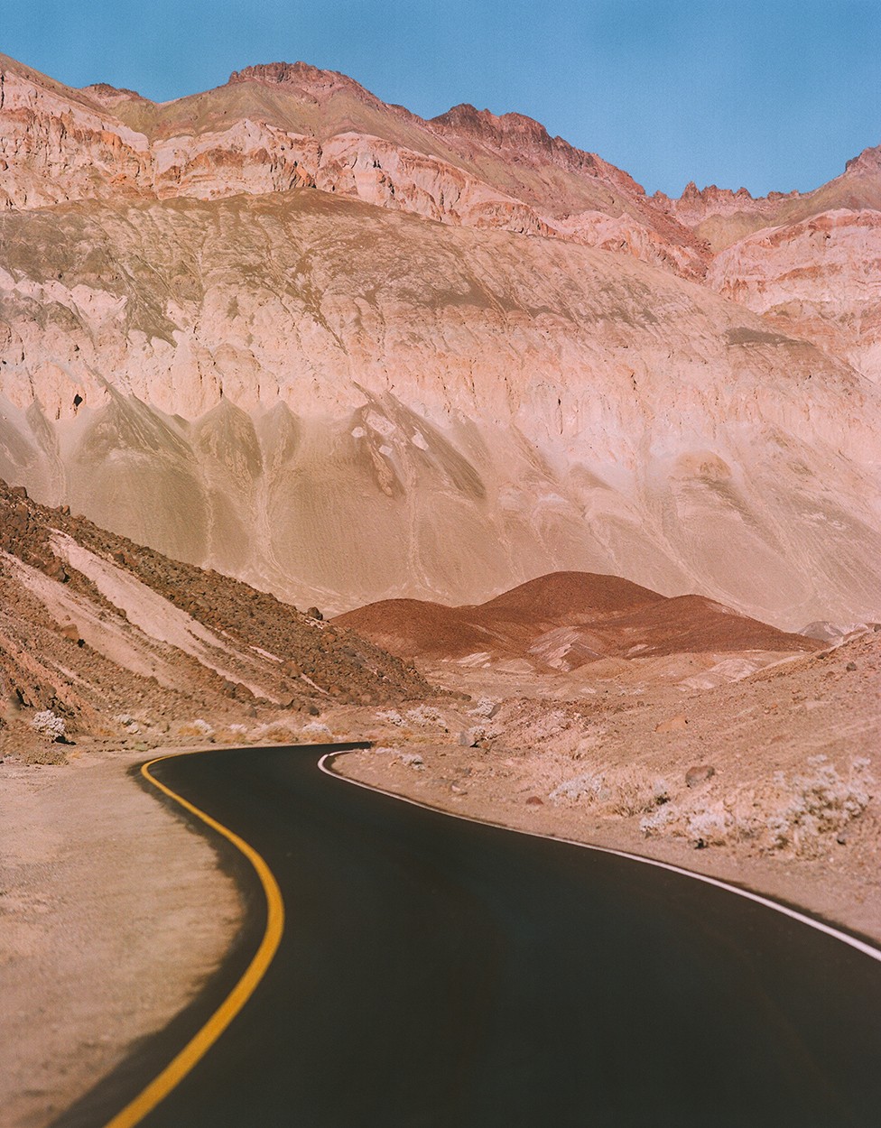 A winding road with a yellow line leads through a desert landscape with large mountains under a clear sky.