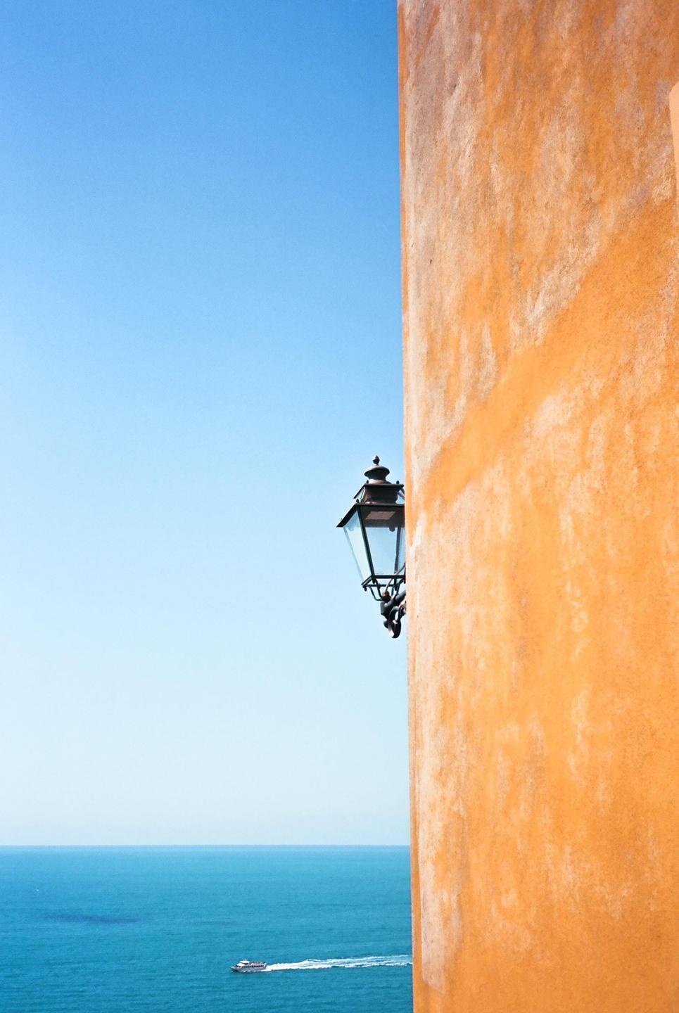 A street lamp is mounted on an orange wall overlooking the sea with a boat in the distance.