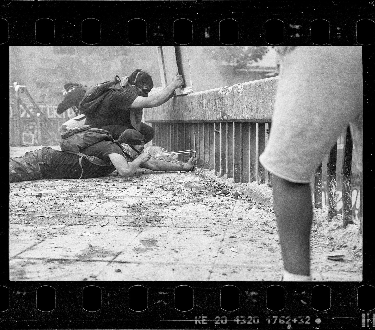 A man lies on the ground aiming a slingshot while another man crouches behind a concrete barrier.