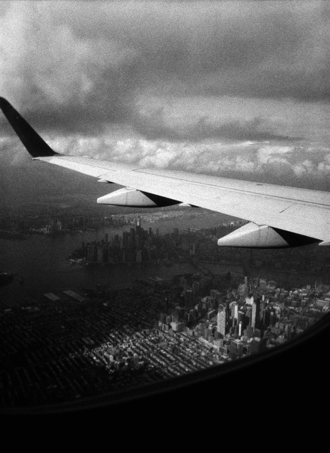 An airplane wing is visible from a window, with a city and clouds below.